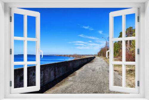 Fototapeta Naklejka Na Ścianę Okno 3D - Asphalt path on the embankment near the water of the lake, sea, river, reservoir and blue distance with the sky with white clouds and horizon. Natural landscape with water on a sunny day