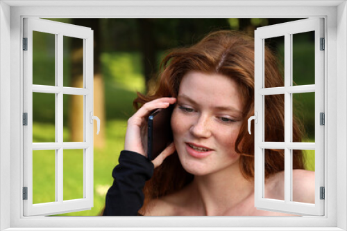 Happy girl with long ginger hair and freckles talking on mobile phone sitting on a bench in summer park