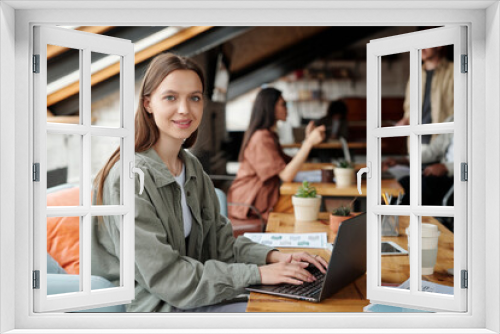 Young smiling female analyst or economist looking at camera while sitting by workplace in front of laptop and working with online financial data