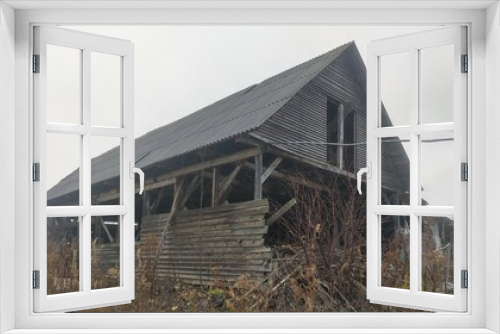 Fototapeta Naklejka Na Ścianę Okno 3D - An old farm building, a barn, stands on a lawn in a rural area. The surrounding area is overgrown with tall grass and bushes. The windows are not glazed and part of the roof is missing. Late autumn