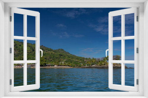 Fototapeta Naklejka Na Ścianę Okno 3D - A picturesque tropical island against a background of blue sky and clouds. Lush green vegetation on the hill. Houses with thatched roofs near the shore. The boats are moored at the water's edge. 