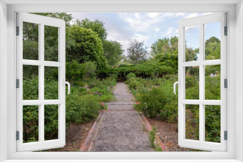 Fototapeta Naklejka Na Ścianę Okno 3D - A gravel walking path lined with bushes and wildflowers leads to an ivy covered entryway in a tranquil garden, with green trees and a partly cloudy sky in the background.