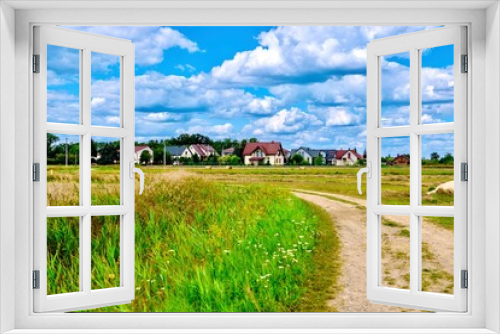 Fototapeta Naklejka Na Ścianę Okno 3D - Country road through the meadow with houses and blue sky with clouds, Poland.