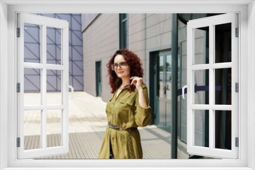 Portrait of a woman standing near a supermarket building. Caucasian model with long brunette hair, wears sunglasses and a khaki dress.