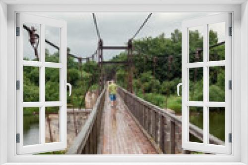 Fototapeta Naklejka Na Ścianę Okno 3D - A person traveling in the distance on a wooden suspension bridge in light summer rain, he is not in a hurry, the beauty and peace of nature