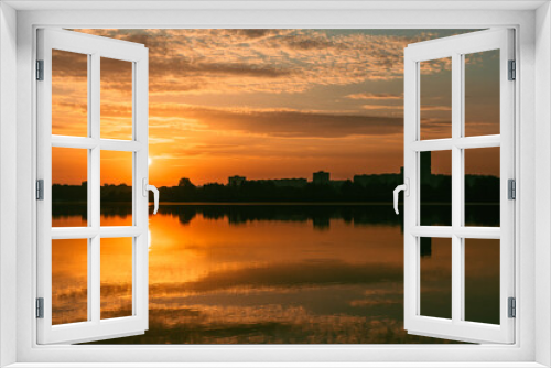 Sunrise sun shining on lake with clouds reflecting in calm mirror water surface. Colorful morning by the river in city park with multistory buildings