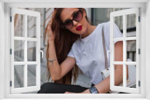 Beautiful fashion urban woman model with cool sunglasses in a white stylish T-shirt and pants with trendy bag sits in the city on the steps