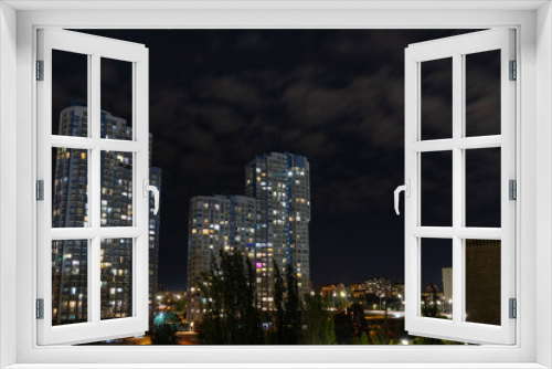 Fototapeta Naklejka Na Ścianę Okno 3D - Evening cityscape with high multi-storey residential buildings. Light from the windows of modern buildings in the city on a summer night