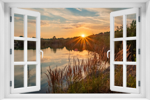 Fototapeta Naklejka Na Ścianę Okno 3D - The image is a splendid showcase of nature's evening adieu, featuring the sun dipping low in the sky, casting its golden glow across a calm lake. Wild grasses in the foreground, kissed by the sunlight