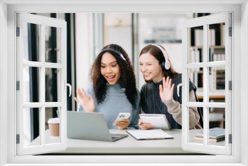 Two Attractive young female college students working on the school project using computer and tablet together, enjoy talking and headphones having a video chat.