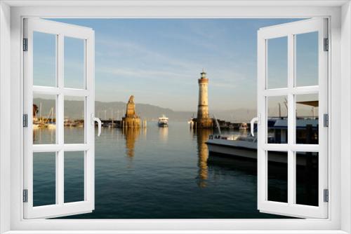 Fototapeta Naklejka Na Ścianę Okno 3D - sunlit harbour and beacon of Lindau on tranquil lake Constance (lake Bodensee) with the Alps in the background on a sunny and warm evening in October (Lindau, Germany)	