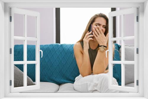 Attractive young hispanic woman's yawn interrupts phone convo while relaxed on sofa at home, seeming tired