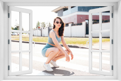 Young beautiful confident female in trendy summer blue cycling shorts and tank top clothes. Carefree woman posing in street. Positive model having fun. Cheerful and happy. Sits on asphalt