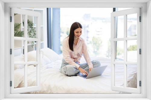 Attractive young woman working remotely from home, typing on modern wireless laptop. Relaxed lady sitting on comfy bed and enjoying online chatting with coworkers on weekend.