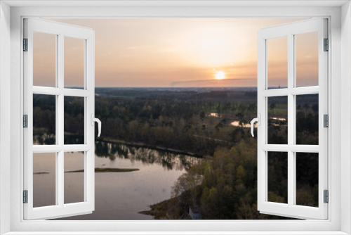 Fototapeta Naklejka Na Ścianę Okno 3D - An aerial view captures the serene beauty of a forest lake at twilight. The setting sun dips towards the horizon, casting a gentle golden glow across the water's surface and the surrounding trees. The