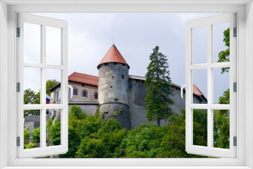 Fototapeta Naklejka Na Ścianę Okno 3D - Scenic view of castle on top of rock with Slovenian flag waving at lakeshore of Slovenian Lake Bled on a cloudy summer day. Photo taken August 8th, 2023, Bled, Slovenia.