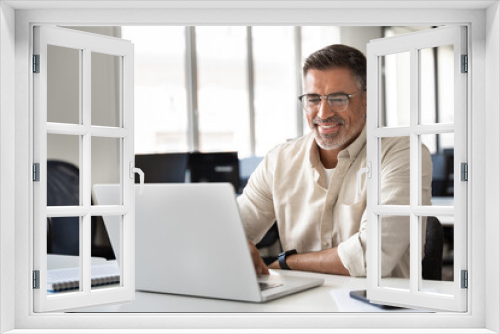 Happy smiling mature Indian or Latin business man ceo trader using computer, typing, working in modern office, doing online data market analysis, thinking planning tech strategy looking at laptop.