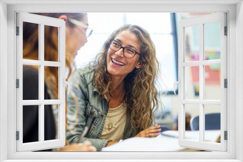 Two women engaged in conversation sitting at desks in a classroom, discussing educational topics and sharing ideas