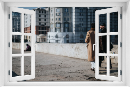 Two fashionably dressed men walk and talk along a city bridge, with blurred urban buildings and a solitary figure in the background.