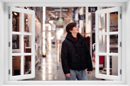 A young woman in casual wear, with a thoughtful expression, browses through a well-lit hardware store aisle, surrounded by shelves of home improvement goods