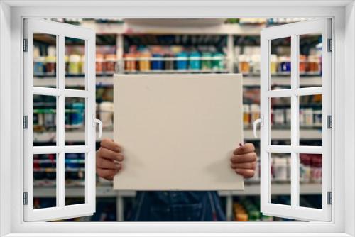 person holding a blank canvas in front of racks of paint