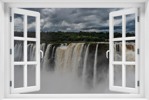 Fototapeta Naklejka Na Ścianę Okno 3D - The incredible powerful waterfall - the Devil's Throat. Streams of foaming white water fall into the abyss. Green vegetation on the river bank. Clouds in the sky. Iguazu Falls. Argentina.