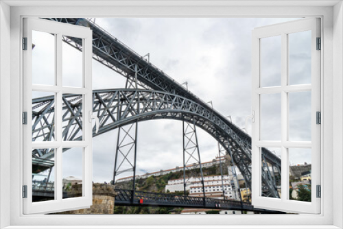 Fototapeta Naklejka Na Ścianę Okno 3D - Low profile view of the Don Luis I steel bridge in Porto with rain clouds in the background and people walking and taking photos of the Douro River on the upper platform and people in restaurants.