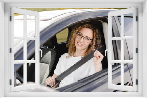 young beautiful smiling woman sitting in motionless car looking straight to camera, fastening her seatbelt, attractive caucasian woman in white t-shirt