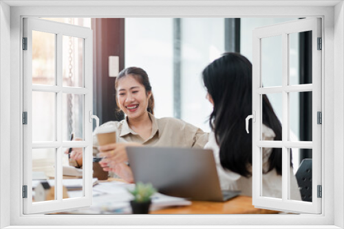 Two joyful women sharing a light-hearted moment with coffee in hand, working together at an office desk.