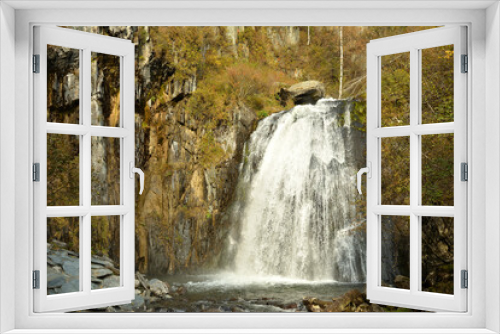 Fototapeta Naklejka Na Ścianę Okno 3D - A powerful waterfall flows in a wide stream from a high mountain, surrounded by yellowed autumn forest.
