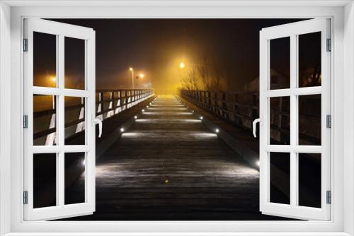 Fototapeta Naklejka Na Ścianę Okno 3D - Dilapidated old vintage pedestrian wooden bridge with wooden handrails renovated and updated with decorative modern LED lights at night engulfed with thick fog and bright city lights in background