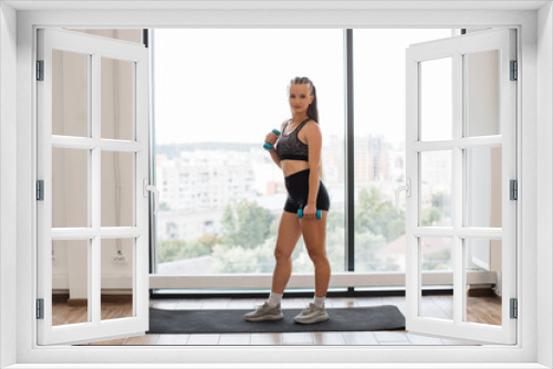 A fit young woman holds blue dumbbells, ready to workout in a bright home environment. She stands on a yoga mat by a large window overlooking the city.