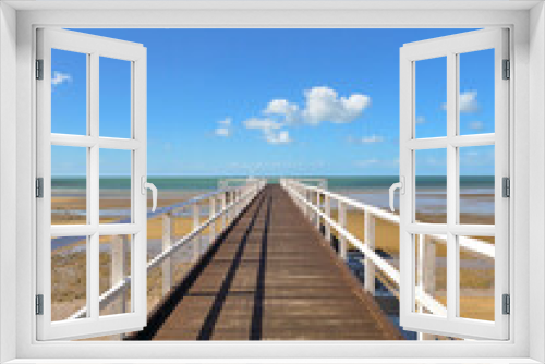 Fototapeta Naklejka Na Ścianę Okno 3D - A long wooden pier stretching to the horizon, over a calm beach at low tide with clear blue sky, yellow sand and little white clouds.