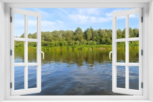Fototapeta Naklejka Na Ścianę Okno 3D - Shrubs and deciduous trees grow on the grassy banks of the river. There are ripples on the water. A heron stands in the water near the opposite bank. Sunny summer weather and blue sky