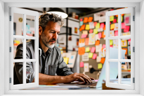 A mature man working on a laptop in a modern office, surrounded by colorful sticky notes. He appears focused and engaged in his task.