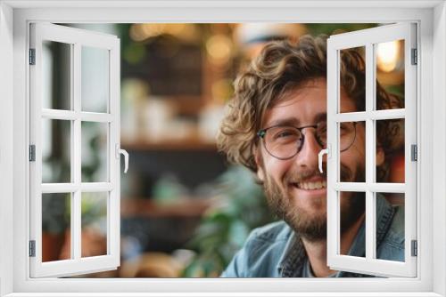 A young man with curly hair and glasses is smiling. He is wearing a blue shirt and a denim jacket. He is standing in a room with plants in the background.