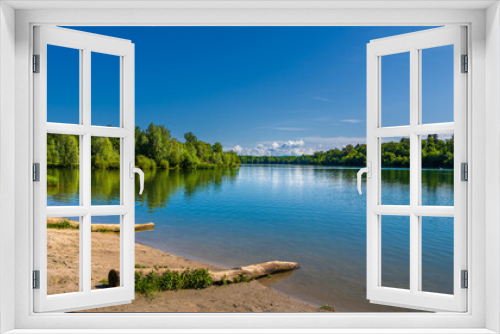 Fototapeta Naklejka Na Ścianę Okno 3D - Shore with logs on water's edge and the blue sky, green trees reflected in the water