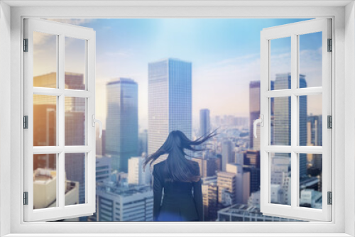 日本の街・都市・朝日を受けたオフィスビル群を前に佇み風を受けて髪をなびかせる会社員の女性の出勤　ビジネス・はじまり・決意・目標・挑戦のイメージ