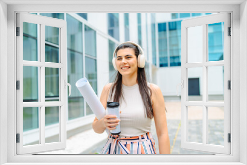 One young caucasian woman student going to university while listening to music on wireless headphones and holding papers and coffee	