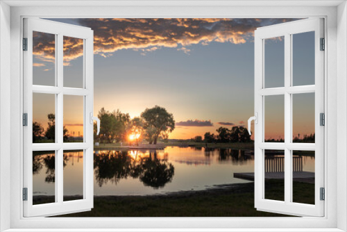Fototapeta Naklejka Na Ścianę Okno 3D - A serene sunset paints the sky with hues of orange and pink, reflecting off the tranquil waters of a park lake. Trees line the shore, creating a picturesque backdrop for a peaceful evening.