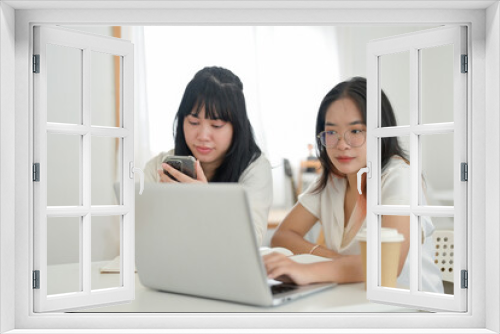 A young Asian woman is working on her laptop computer while her friend is using a smartphone.