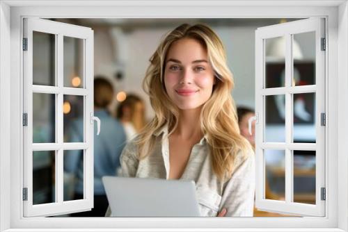 A smiling young woman with blonde hair holding a laptop in an office environment, exuding confidence and professionalism