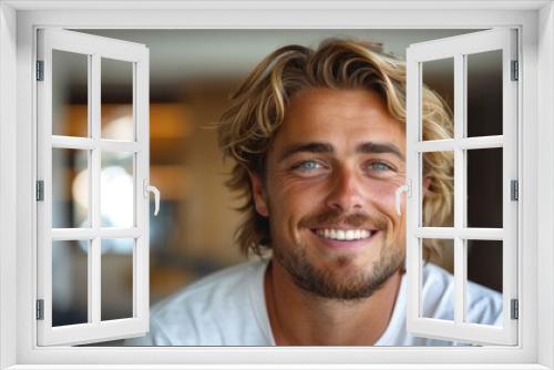 A cheerful man with blonde hair, smiling warmly at the camera, presenting an inviting and joyful expression, captured in a bright and modern indoor environment.