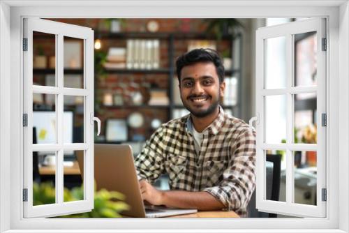 A man is sitting at a desk with a laptop in front of him. He is smiling and he is happy