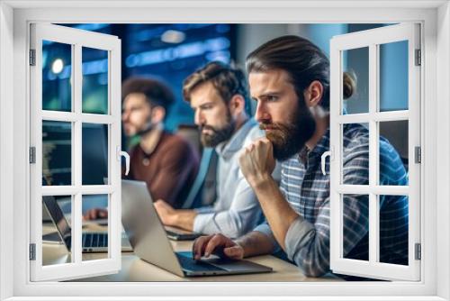 Sofware developer thinking while touching beard while typing on laptop sitting at desk with multiple screens parsing code. Focused database admin working with team coding in the background.