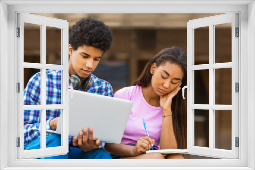 A teen black guy sits outside, using a laptop while a teen girl takes notes in a notebook. They appear to be engaged in a learning or studying activity, perhaps a school project