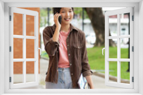 A high school student walks along a red brick school building, talking on her phone. She holds books and a tablet in her other hand