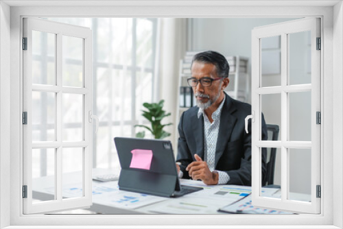 Mature businessman immersed in work at his office desk, focused on his laptop in a bright, modern workplace flooded with natural light