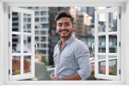 A young man with a cheerful demeanor is smiling confidently while standing on a rooftop with a laptop, with a modern cityscape forming the background.