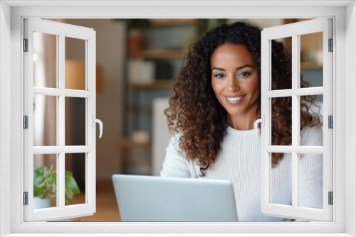 A woman with curly hair and a warm smile is working on her laptop in a comfortable and well-lit living room, surrounded by indoor plants and cozy decor.